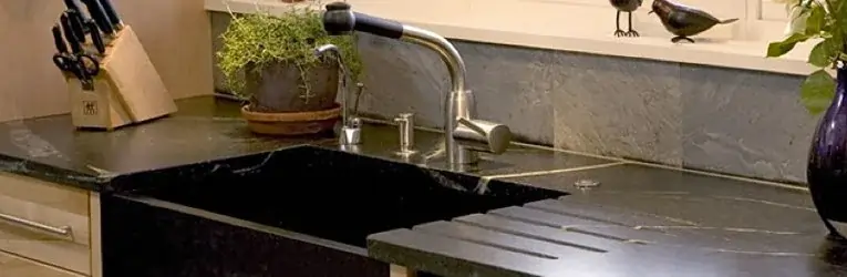 Kitchen with dark soapstone countertop and integrated sink, stainless steel faucet, and natural stone backsplash.
